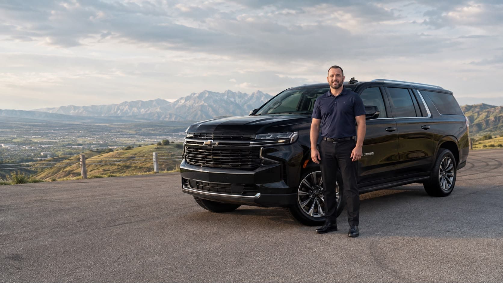 A PrimeKar driver in a navy polo standing beside a black Chevrolet Suburban with a mountain valley behind him